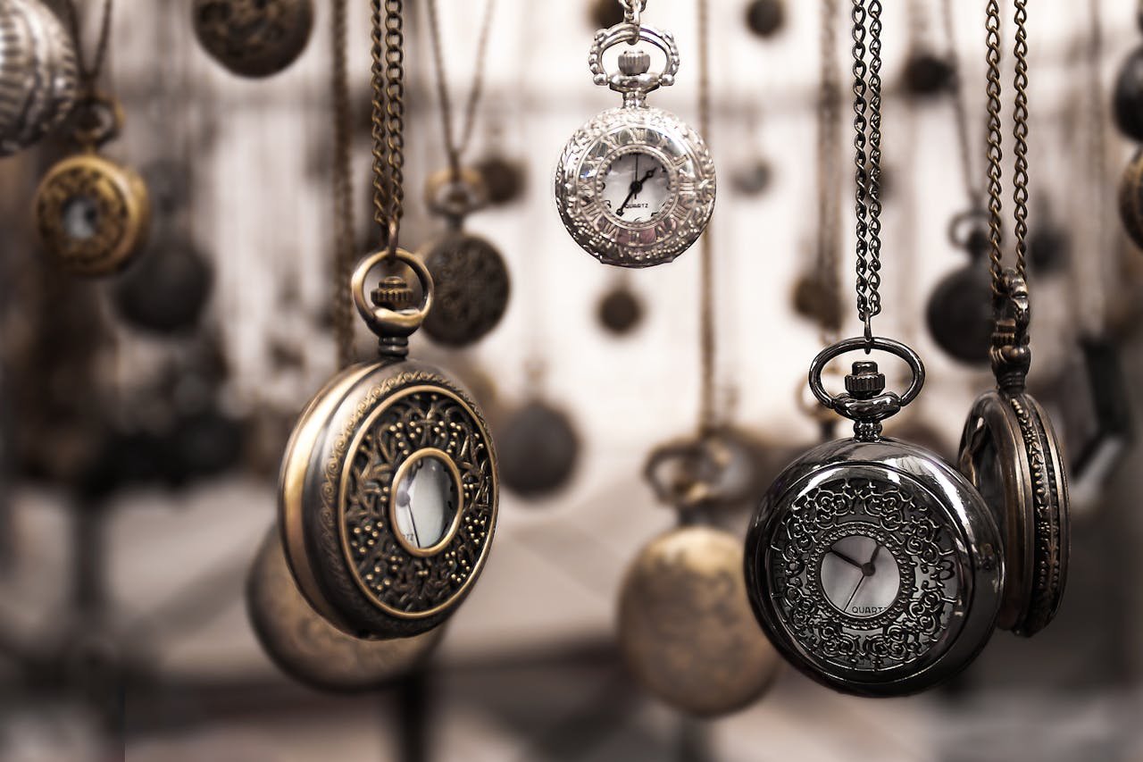 about-us Close-up of antique pocket watches with intricate designs hanging in a blurred vintage setting.